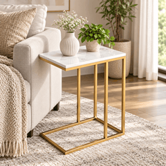 Gold frame C shaped side table with a white marble look top beside a cream sofa holding a flower vase and a plant