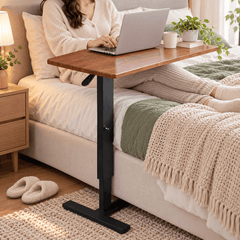 Woman using a laptop on a height adjustable table with a wood look top over a bed, with a mug, a plant and a book on it