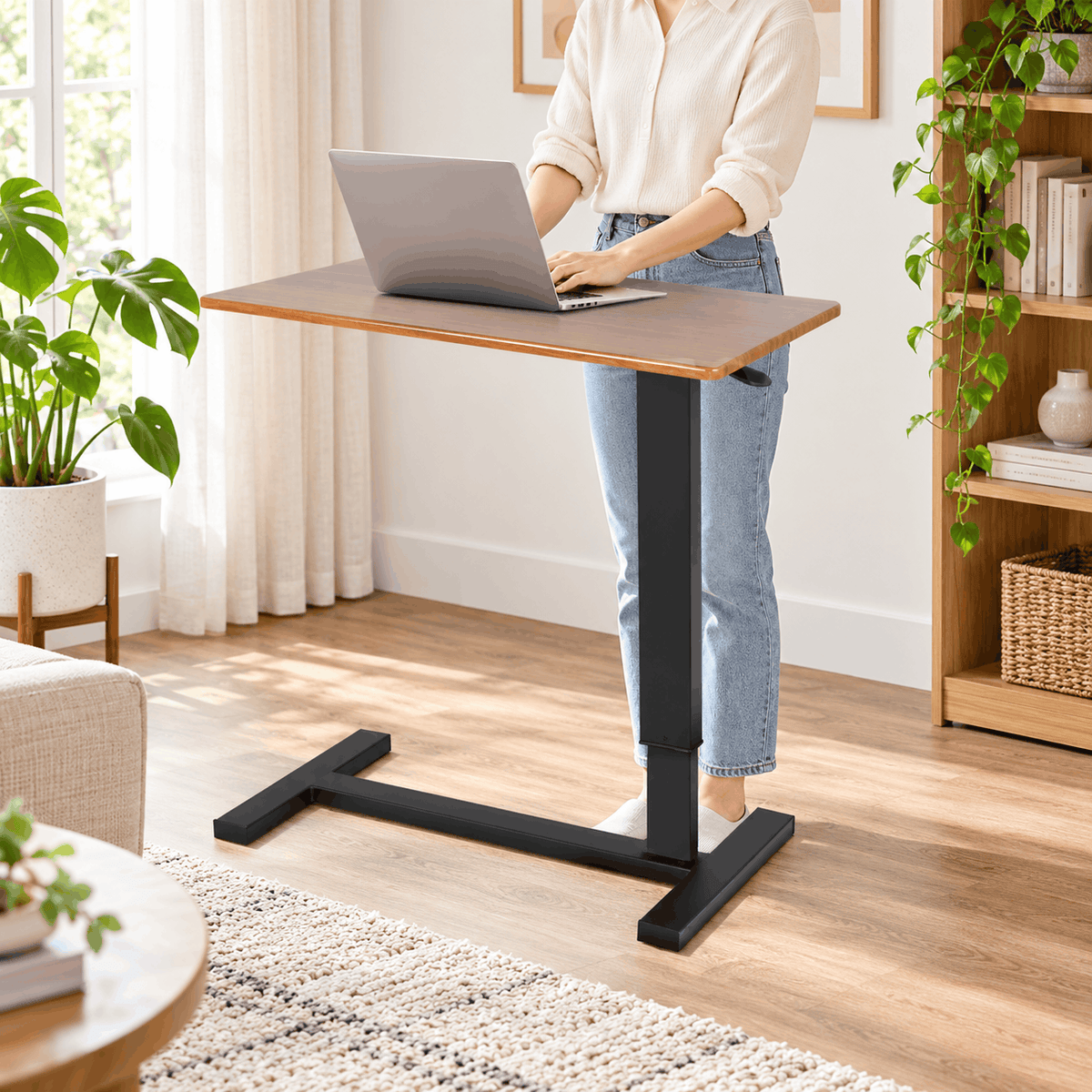 Person using a laptop at a raised height adjustable table with a wood look top and black base in a bright living room