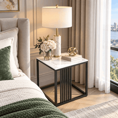 Square side table with a white marble look top and black frame beside a bed holding a table lamp, flowers and books