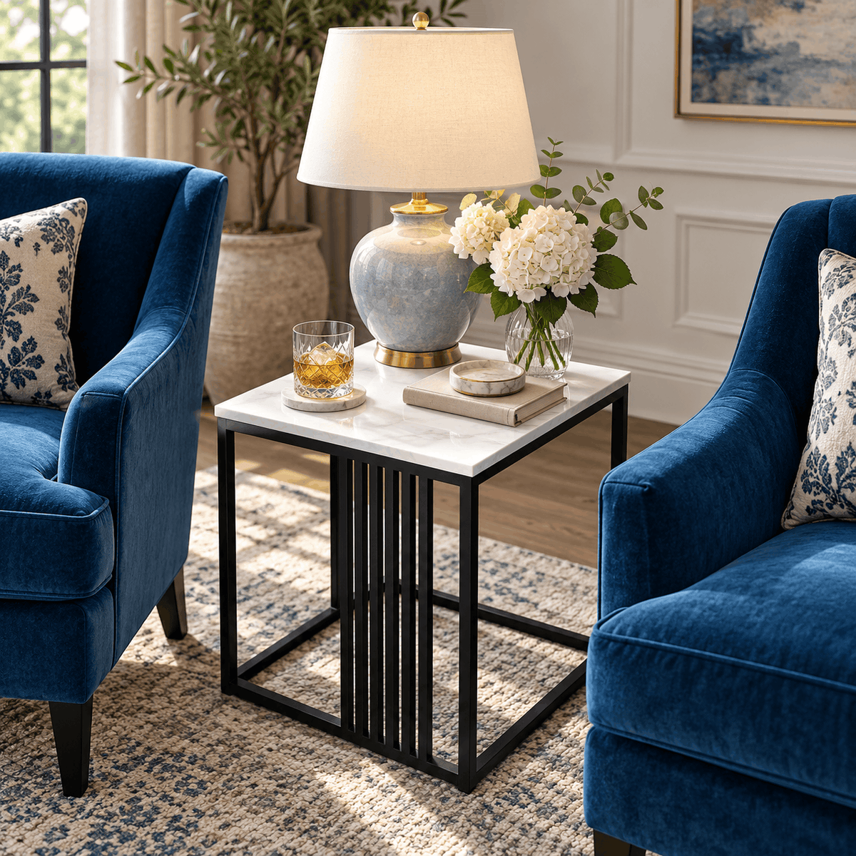 Square marble look side table with a black frame between two blue armchairs holding a table lamp, flowers and a glass