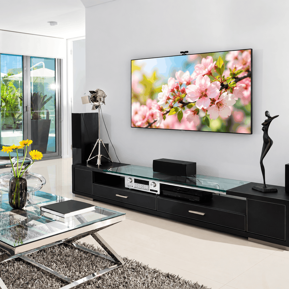 Wall mounted TV above a black TV unit with speakers and an amplifier in a bright living room with glass doors